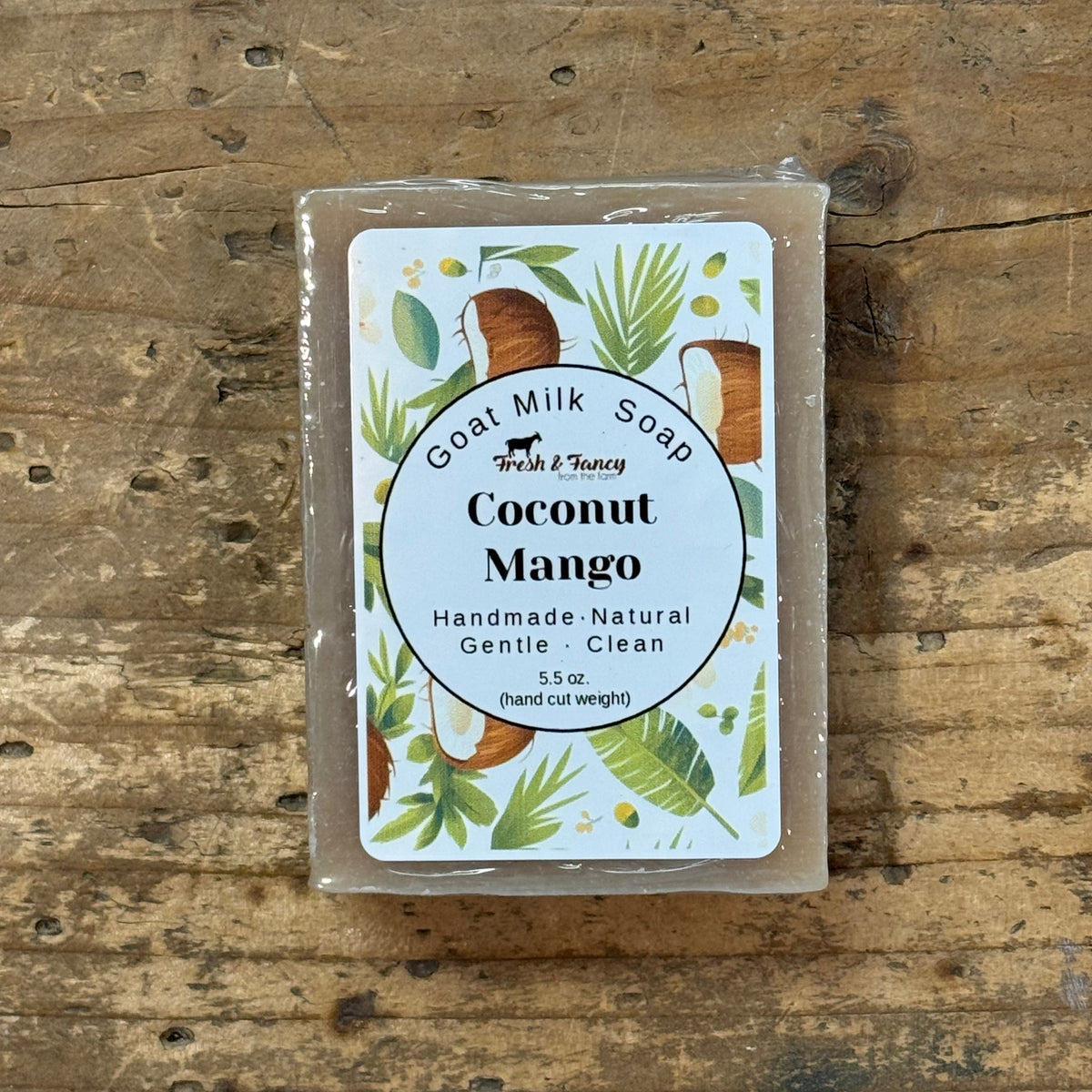 A rectangular bar of goat milk soap in a clear plastic wrap, featuring a white label with 'Coconut Mango' and tropical illustrations, placed on a rustic wooden surface.