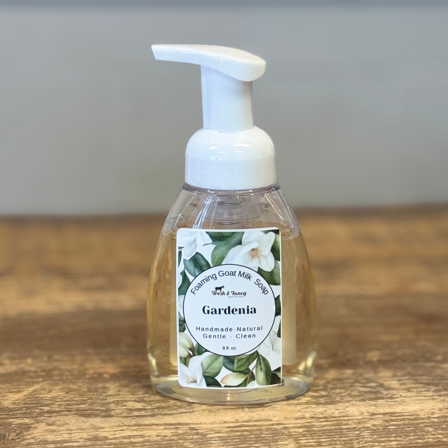 A clear plastic pump bottle of Gardenia handless soap with a white foam pump, sitting on a rustic wooden tabletop with a blurred gray background.