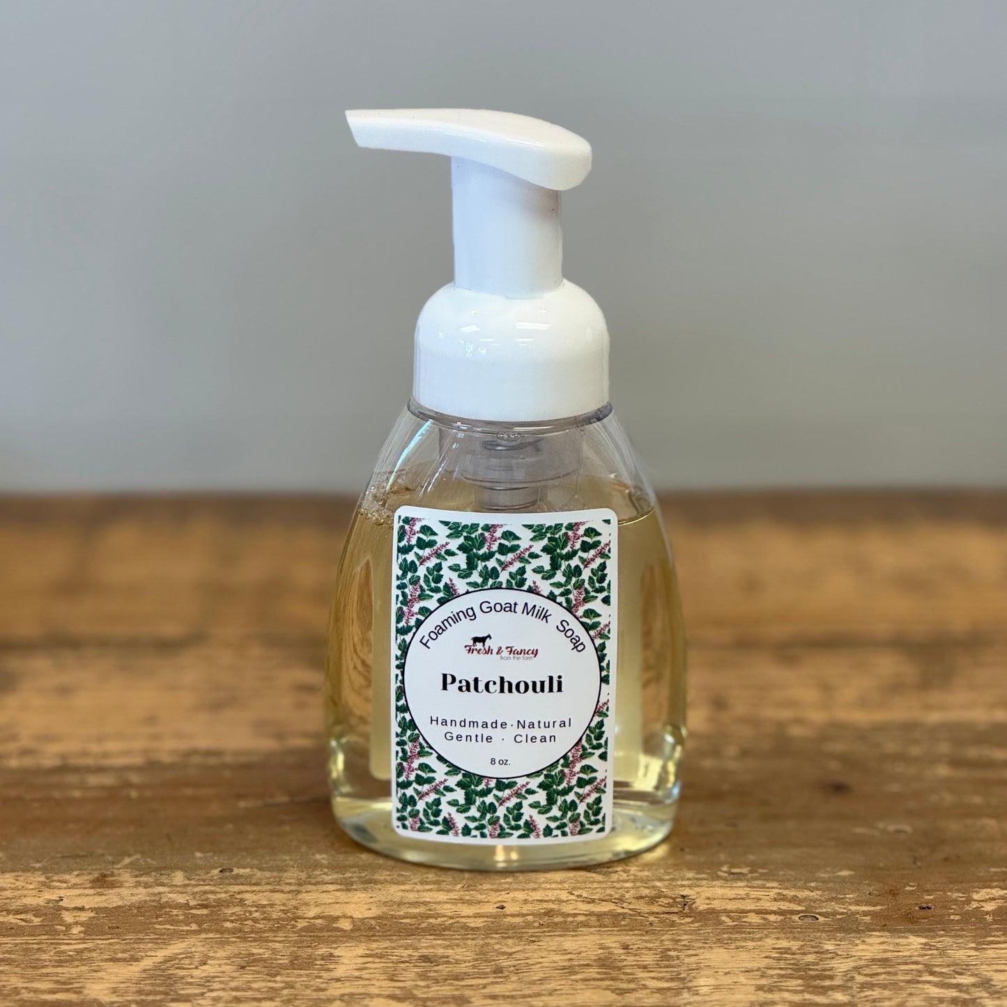A clear plastic pump bottle of Patchouli hand soap with a white foam pump, sitting on a rustic wooden tabletop against a neutral gray background.