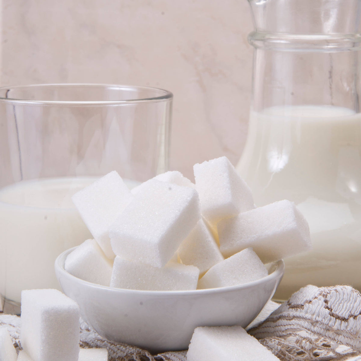 White sugar cubes in a bowl with a glass of milk and a jar on a textured surface.
