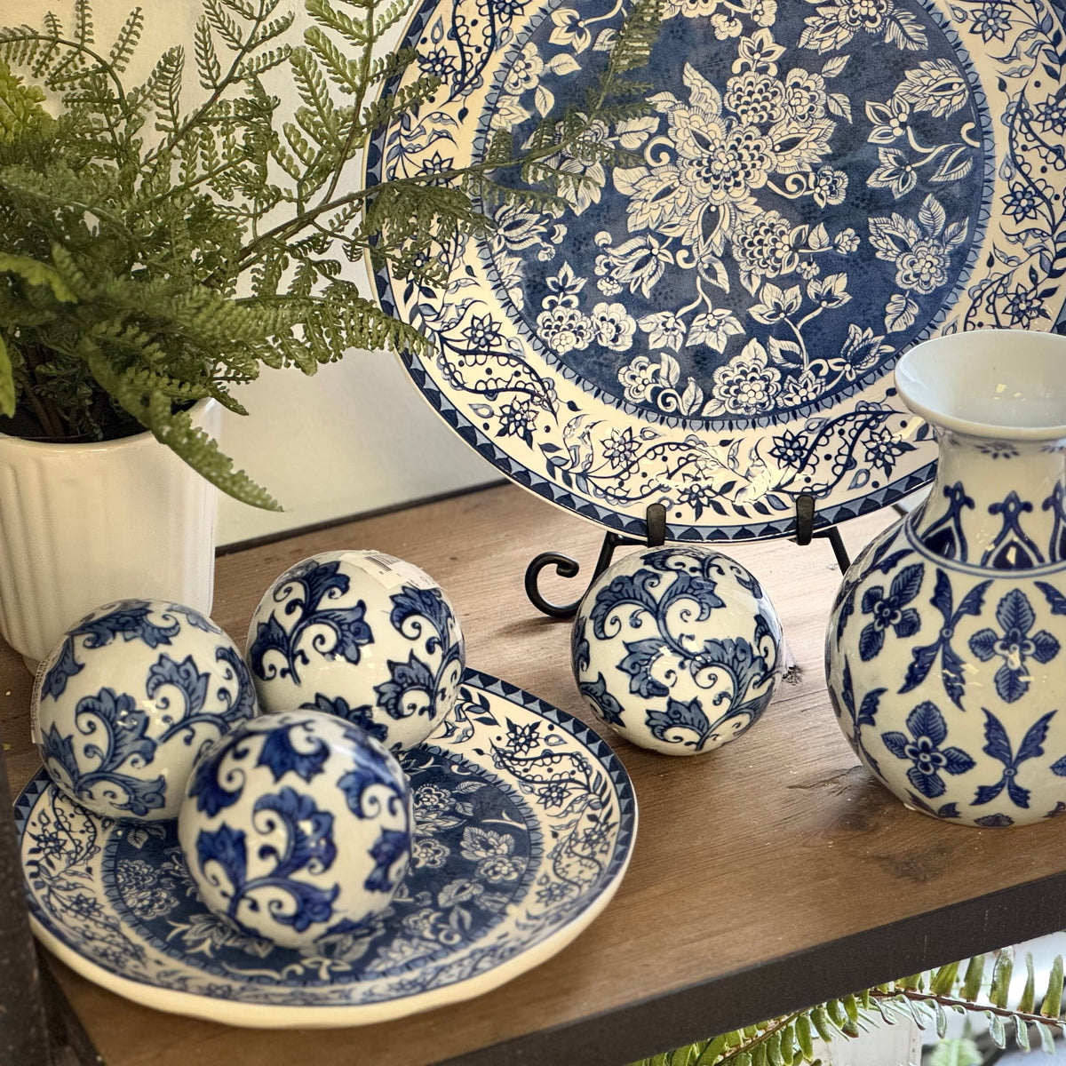 Blue and white floral porcelain tableware set including a vase, plates, and decorative balls, displayed on a wooden shelf with a potted fern.