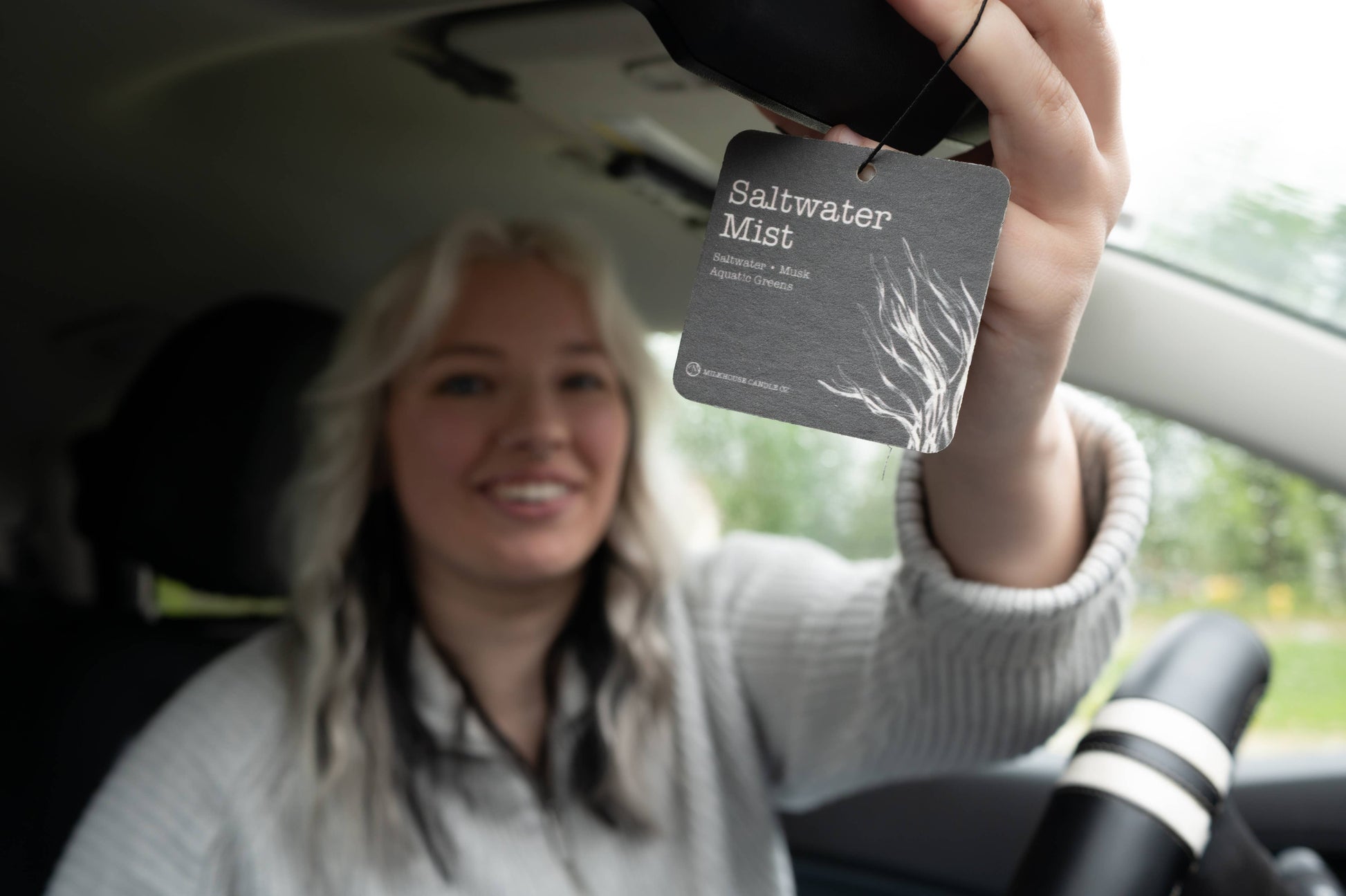 Person holding a car air freshener labeled 'Saltwater Mist' inside a vehicle.