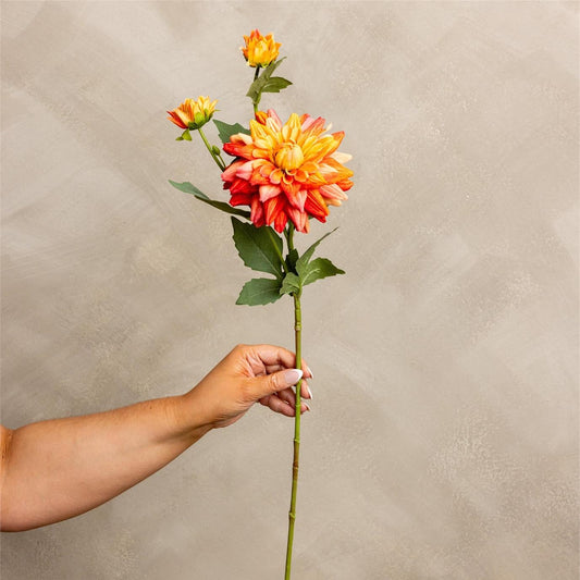 A hand holding a long-stemmed artificial dahlia flower with orange, yellow, and red petals and green leaves, set against a textured beige background.