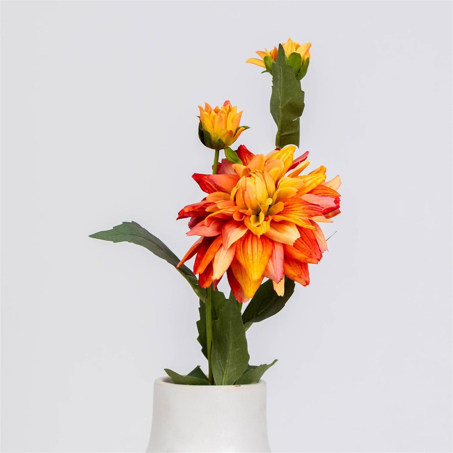 A single artificial-looking dahlia stem with three blooms in orange, yellow, and red tones, green leaves, and a small white vase against a light gray studio background.