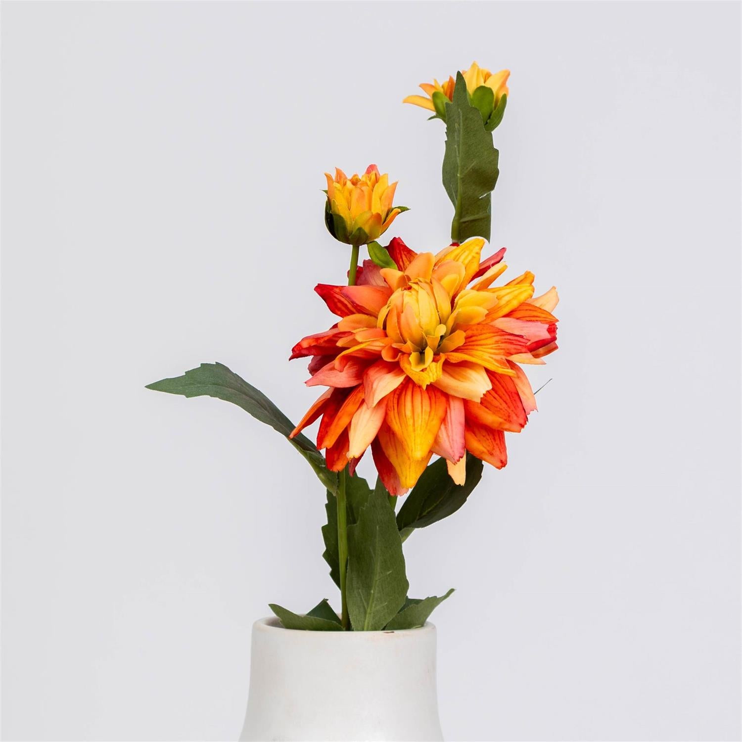 A single artificial-looking dahlia stem with three blooms in orange, yellow, and red tones, green leaves, and a small white vase against a light gray studio background.