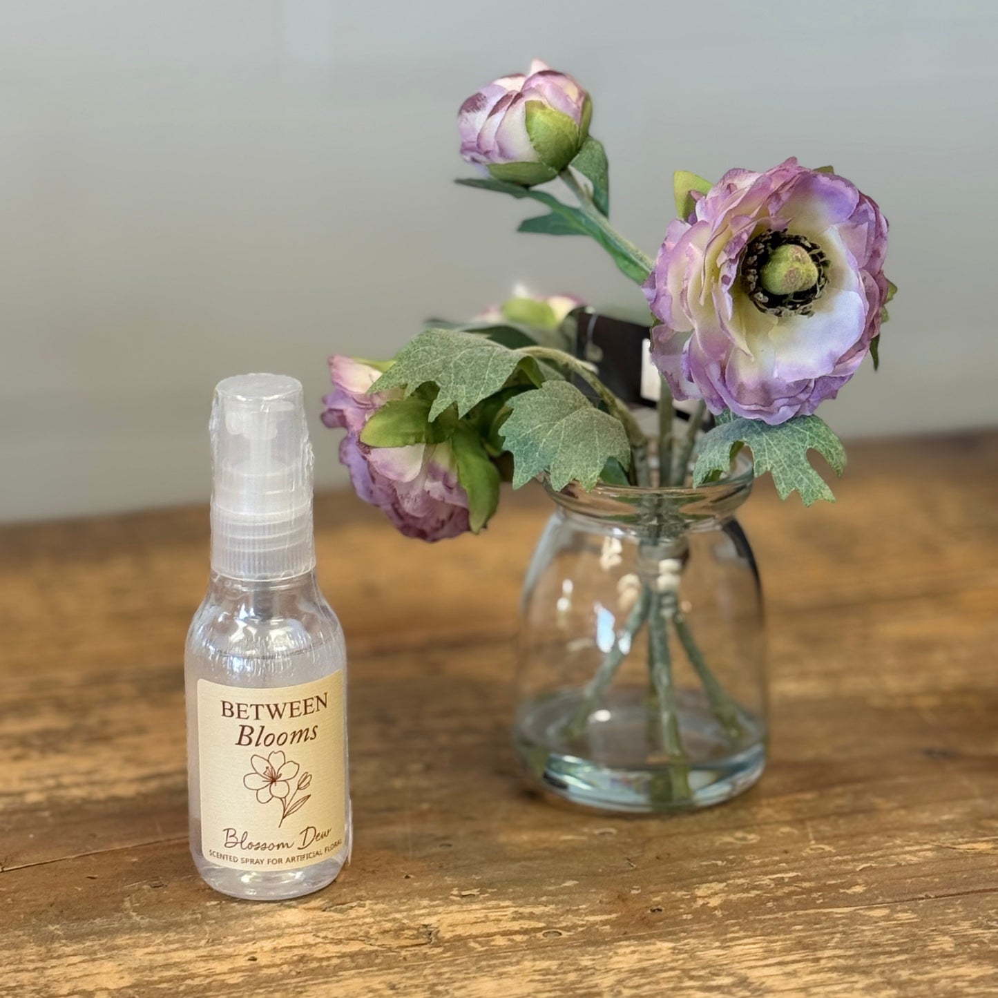 A small clear spray bottle labeled 'Between Blooms' stands on a rustic wooden table beside a clear glass jar holding purple ranunculus flowers.