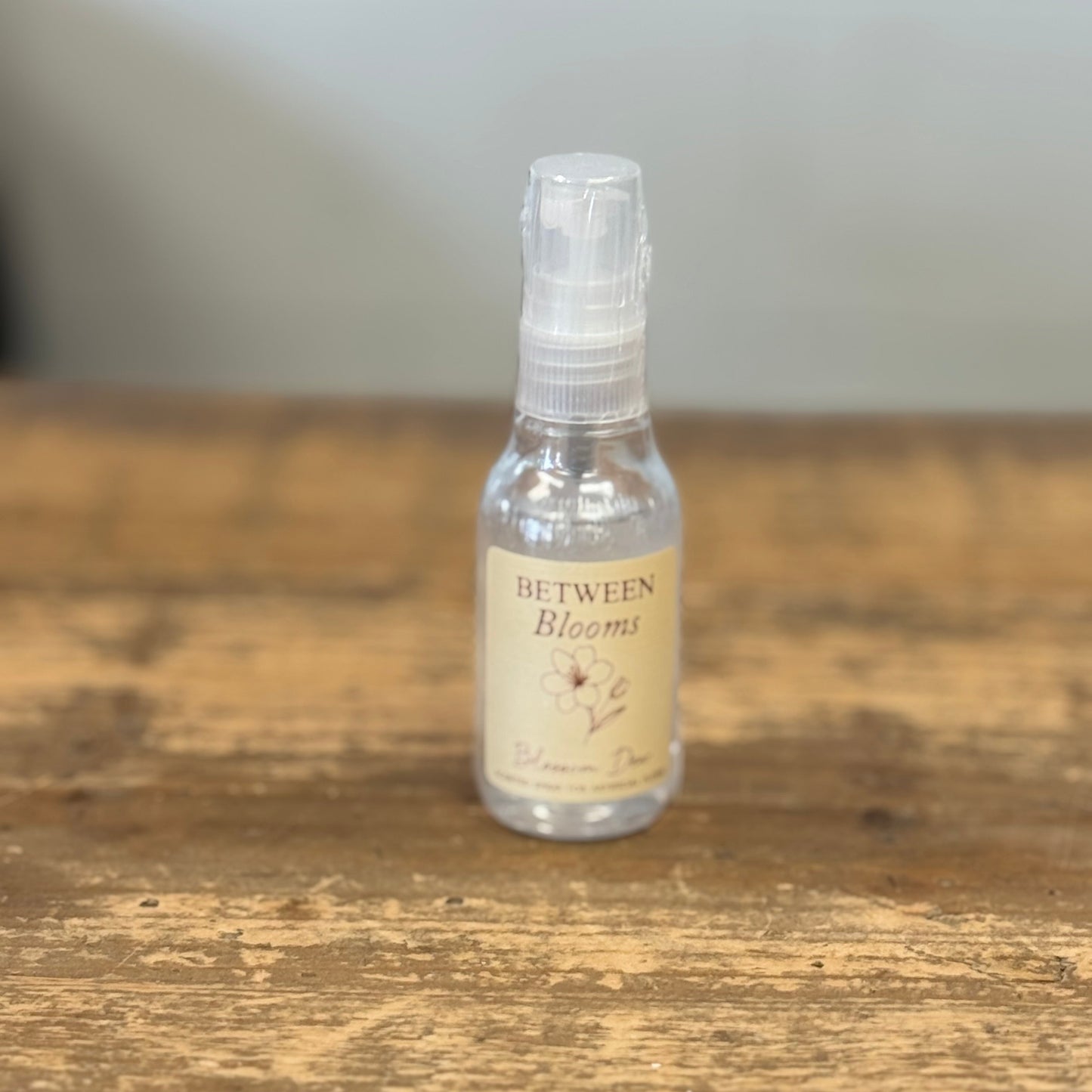 A small clear spray bottle with a beige label reading 'Between Blooms' sits on a rustic wooden tabletop against a soft gray background.