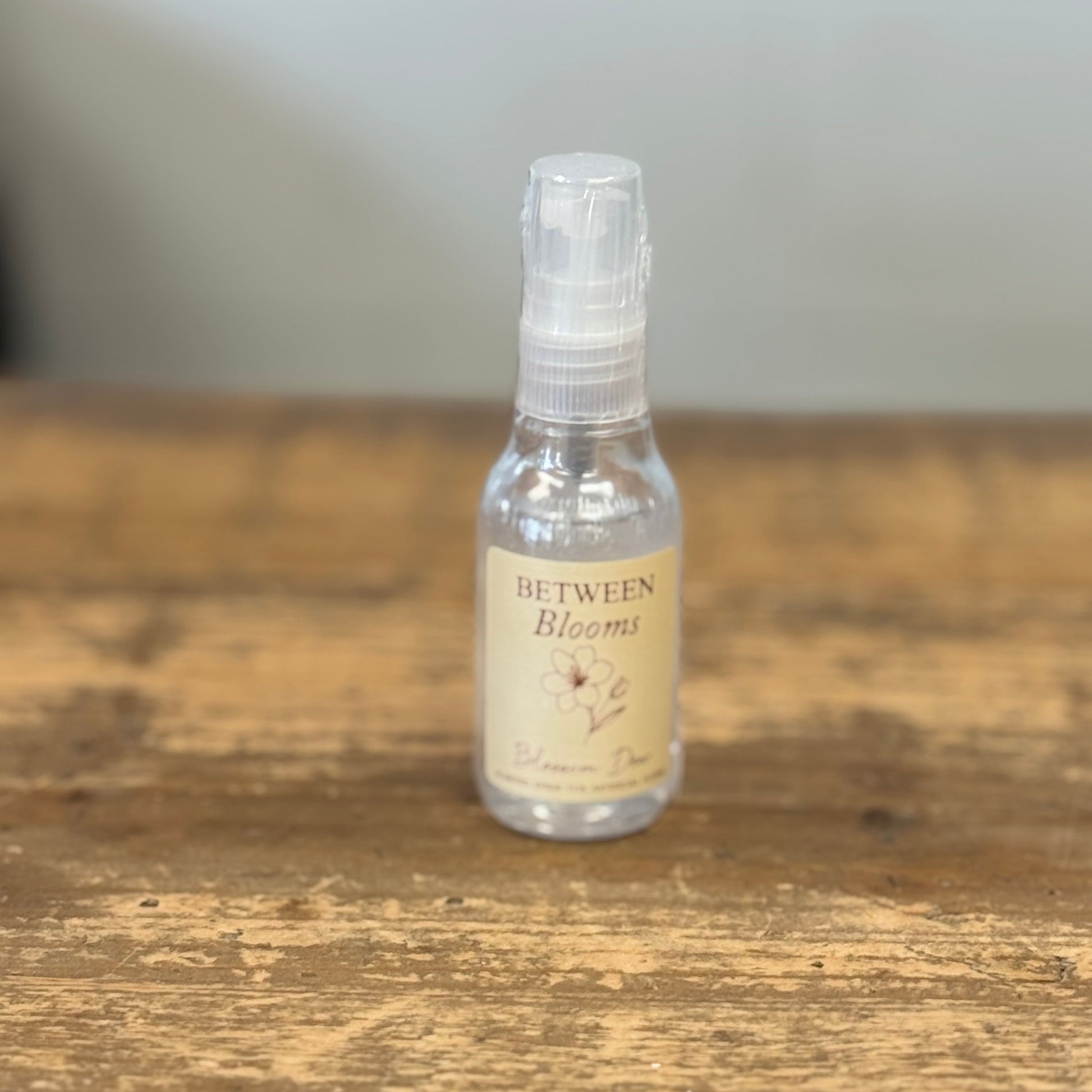 A small clear spray bottle with a beige label reading 'Between Blooms' sits on a rustic wooden tabletop against a soft gray background.