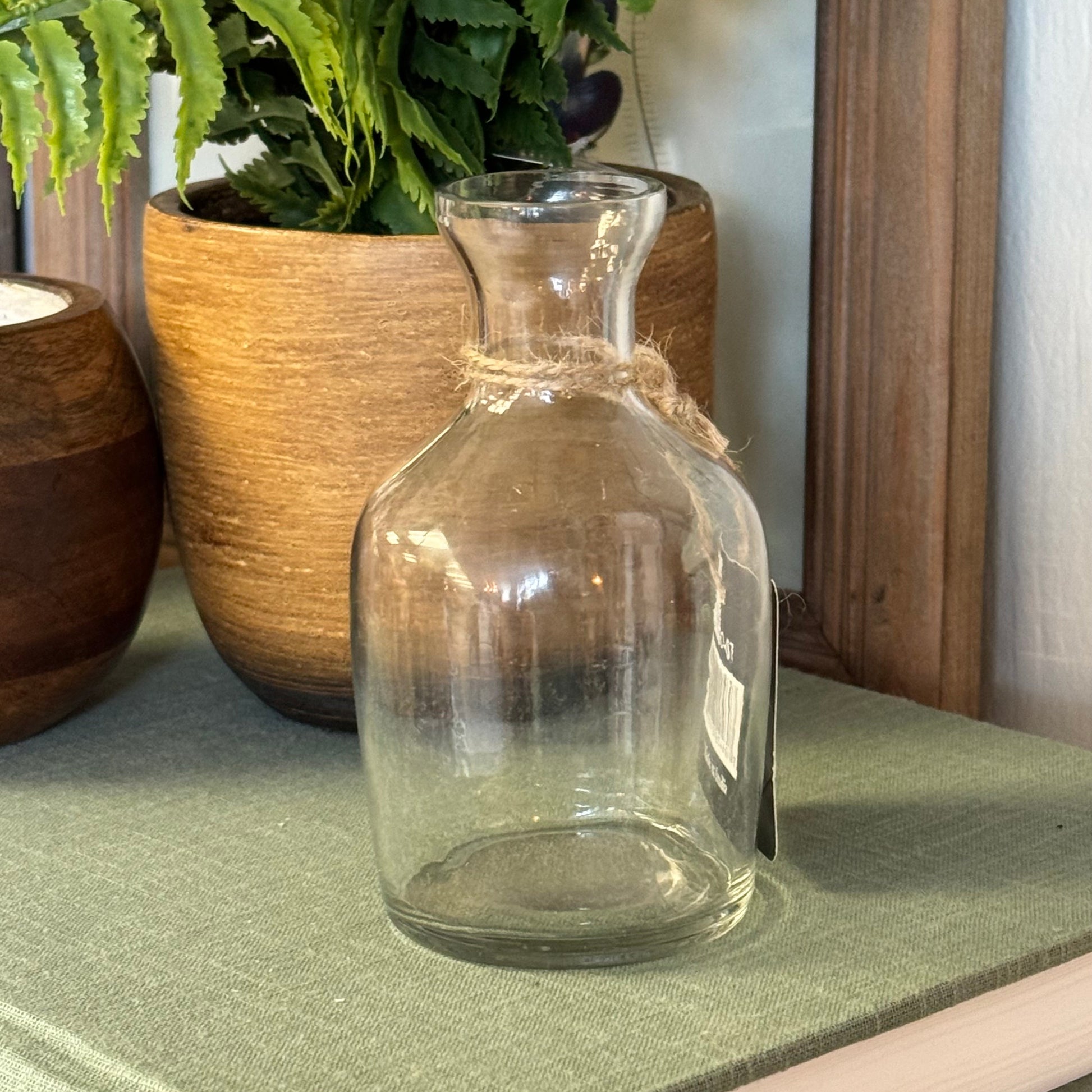 A clear glass bottle vase with a short neck and a rustic twine bow, displayed on a green book next to potted green plants.