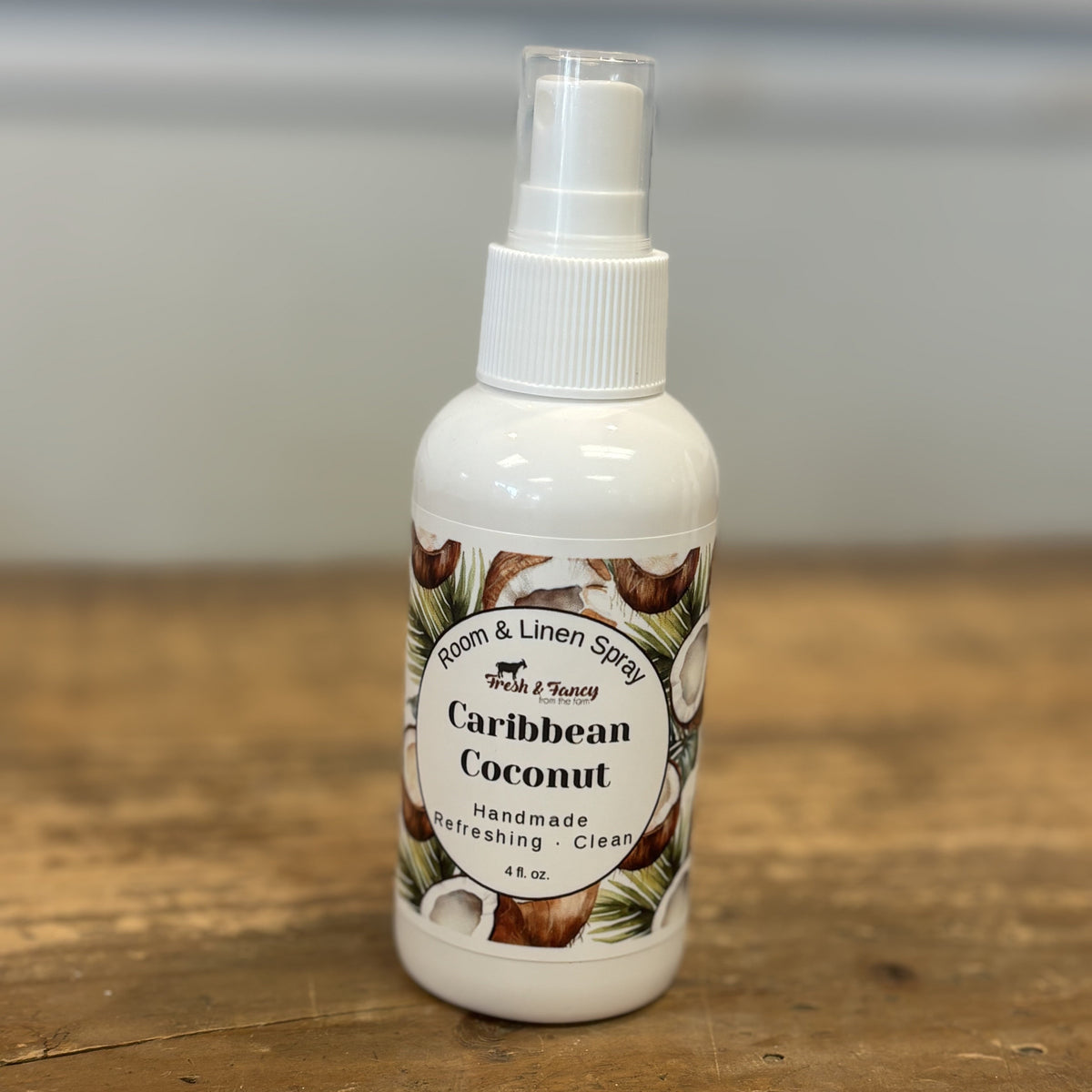 A white plastic spray bottle of Spice & Scents 'Caribbean Coconut' room and linen spray, resting on a rustic wooden tabletop with a blurred gray background.