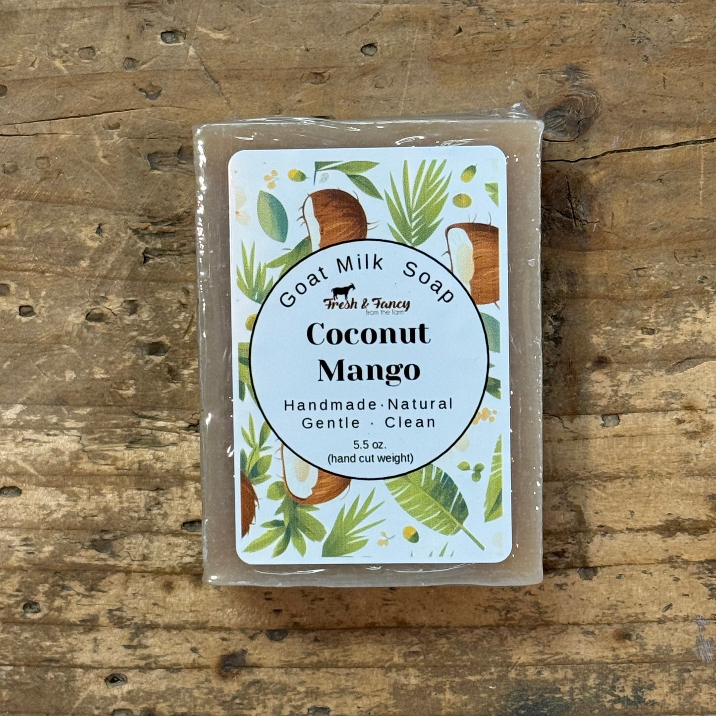 A rectangular bar of goat milk soap in a clear plastic wrap, featuring a white label with 'Coconut Mango' and tropical illustrations, placed on a rustic wooden surface.