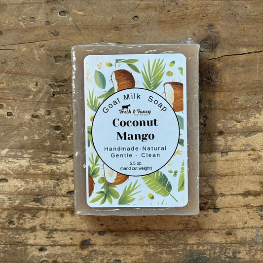 A rectangular bar of goat milk soap in a clear plastic wrap, featuring a white label with 'Coconut Mango' and tropical illustrations, placed on a rustic wooden surface.