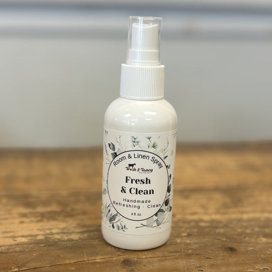 A white plastic room and linen spray bottle with a clear spray top, standing on a rustic wooden tabletop. The label reads 'Fresh & Clean' and features a circular floral design.