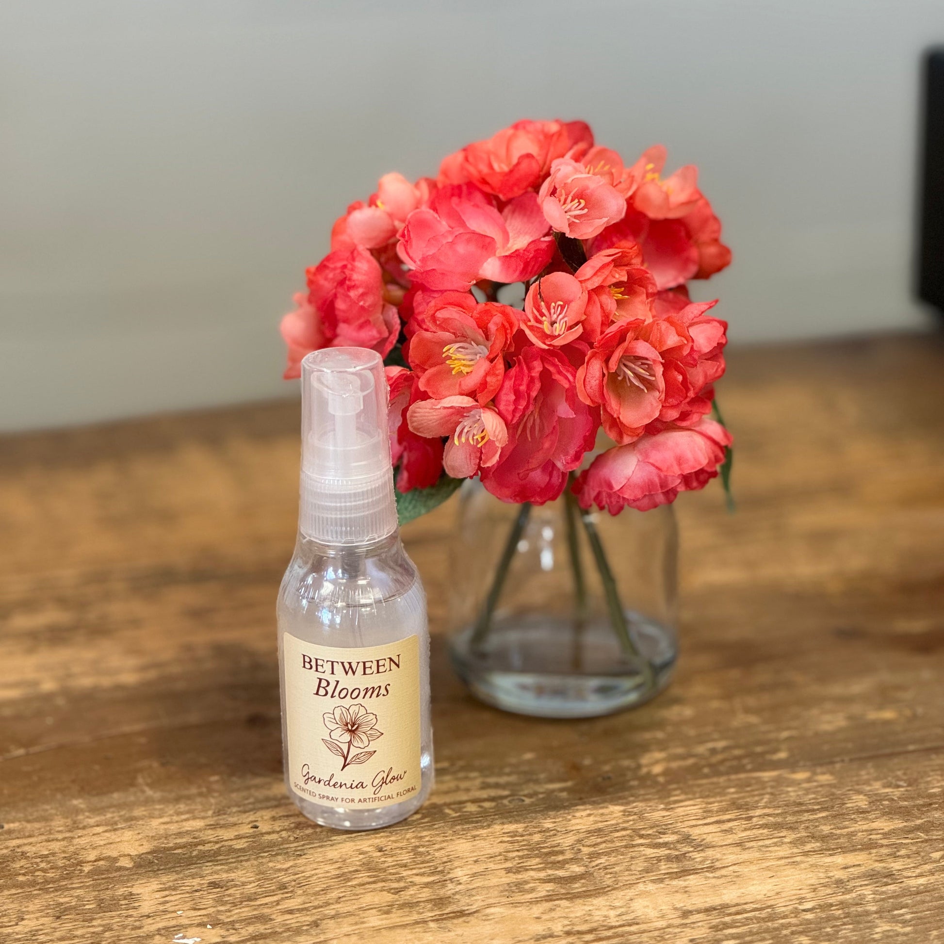 A small clear spray bottle labeled 'Between Blooms' sits on a wooden table in front of a glass vase filled with bright coral-pink flowers.