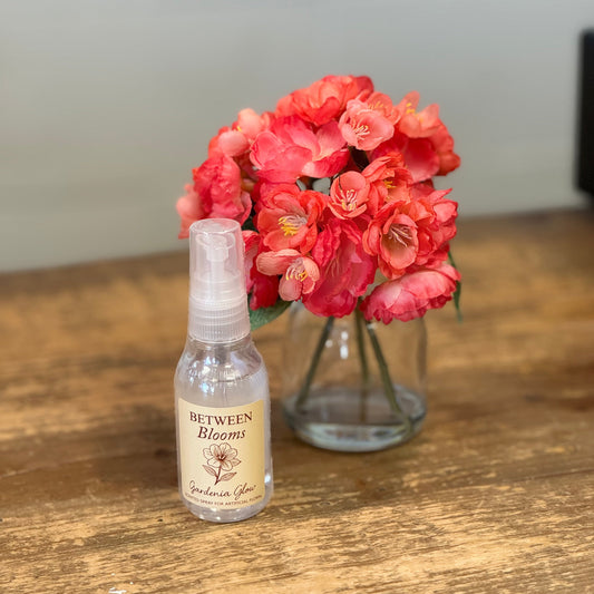 A small clear spray bottle labeled 'Between Blooms' sits on a wooden table in front of a glass vase filled with bright coral-pink flowers.