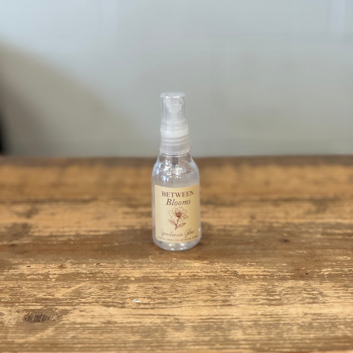 A small clear spray bottle with a beige label reading 'Between Blossoms' sits on a rustic wooden tabletop against a softly blurred light gray background.