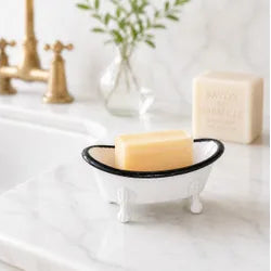 A small white bathtub-shaped soap dish with a black rim holding a bar of soap, placed on a white marble bathroom counter with a gold faucet and a vase of greenery in the background.