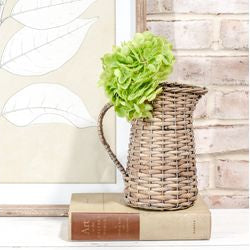 A woven wicker pitcher vase holding a light green hydrangea bloom, styled on top of a vintage book against a brick wall and framed leaf print background.