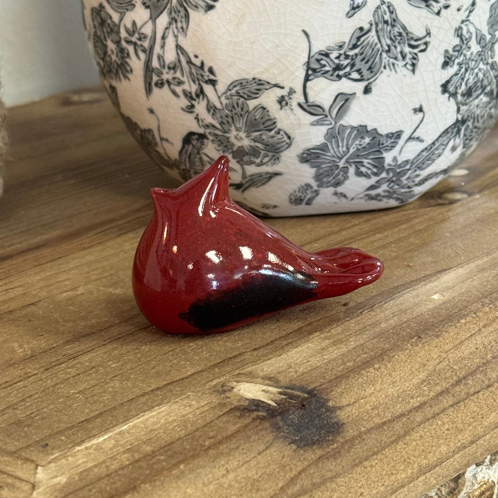 A glossy red ceramic bird figurine, likely a cardinal, resting on a rustic wooden surface with a black and white floral patterned vase in the background.