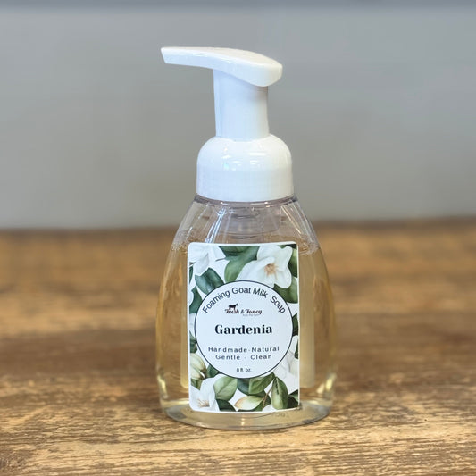A clear plastic pump bottle of Gardenia handless soap with a white foam pump, sitting on a rustic wooden tabletop with a blurred gray background.