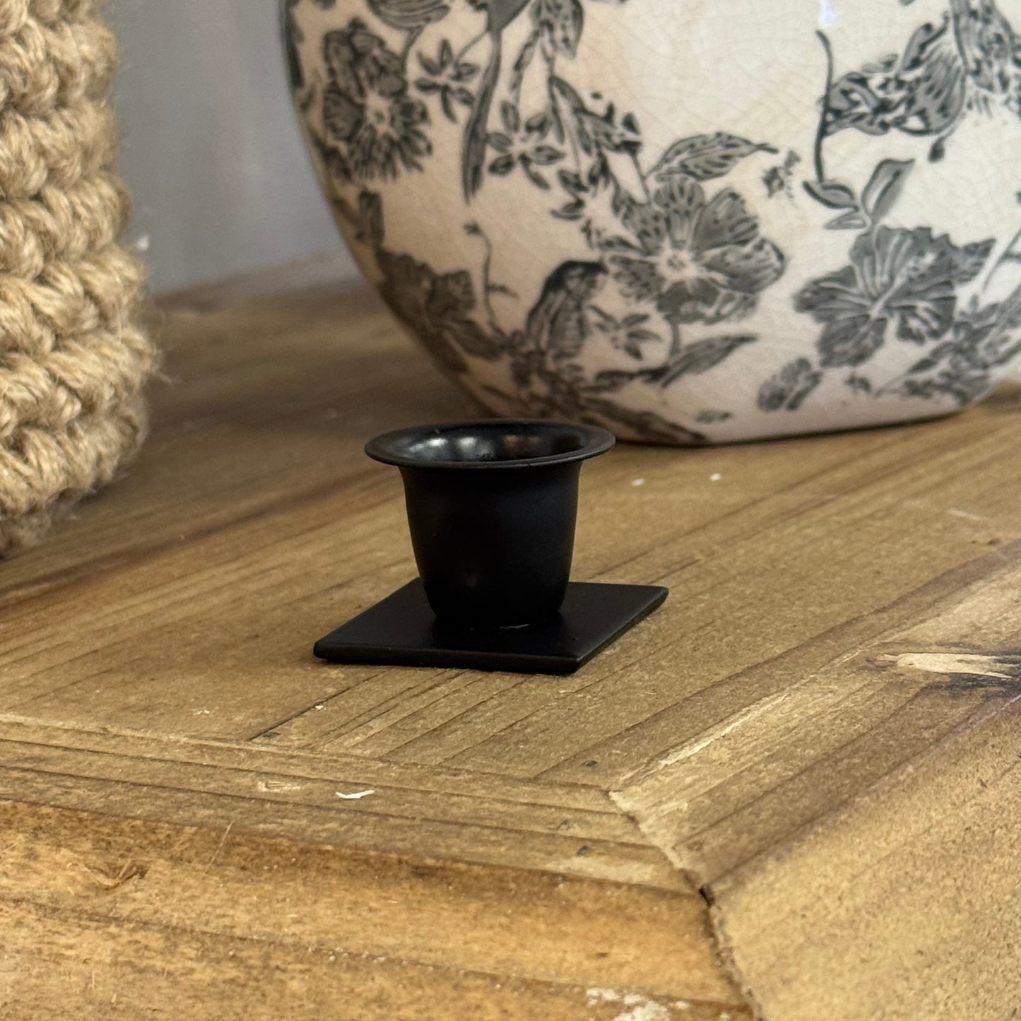 A small matte black metal candle holder with a square base and cup-shaped top, displayed on a rustic wooden mantel next to a patterned ceramic vase.
