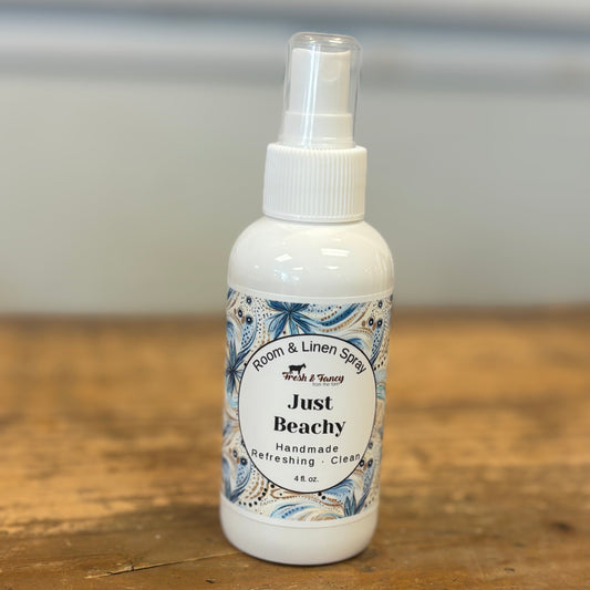 A small white spray bottle labeled 'Just Beachy' room and linen spray, placed on a rustic wooden tabletop with a soft gray background.
