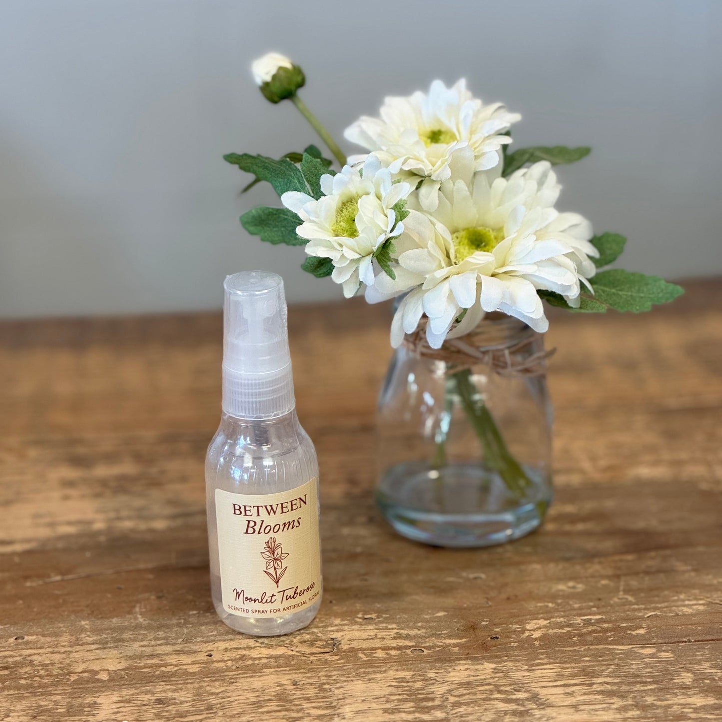 A small clear spray bottle labeled 'Between Blooms' stands on a rustic wooden table beside a glass jar holding white daisies with green leaves, against a soft gray wall.