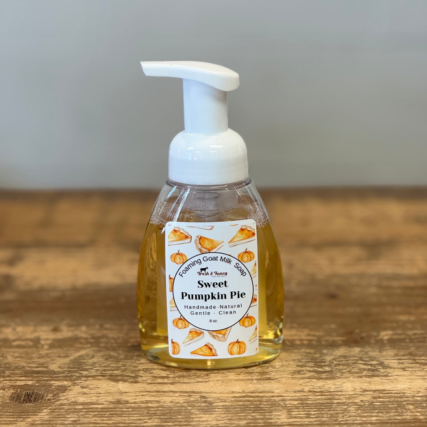 A bottle of Sweet Pumpkin Pie hand foam with a white pump dispenser and a white label decorated with pumpkin illustrations, sitting on a wooden tabletop.