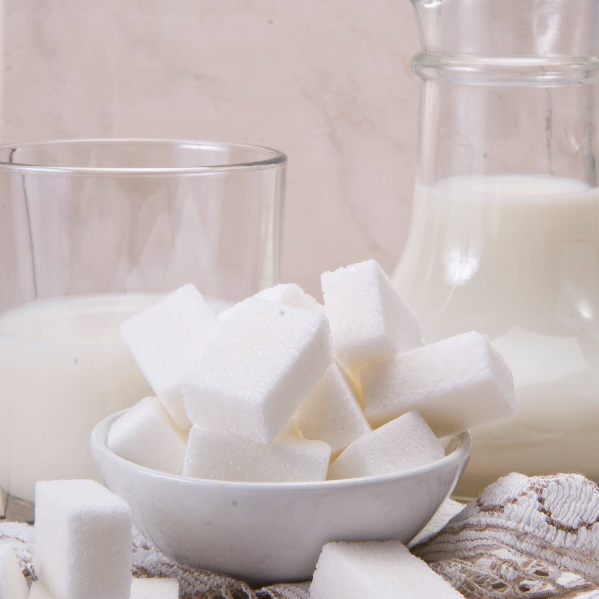 White sugar cubes in a bowl with a glass of milk and a jar on a textured surface.