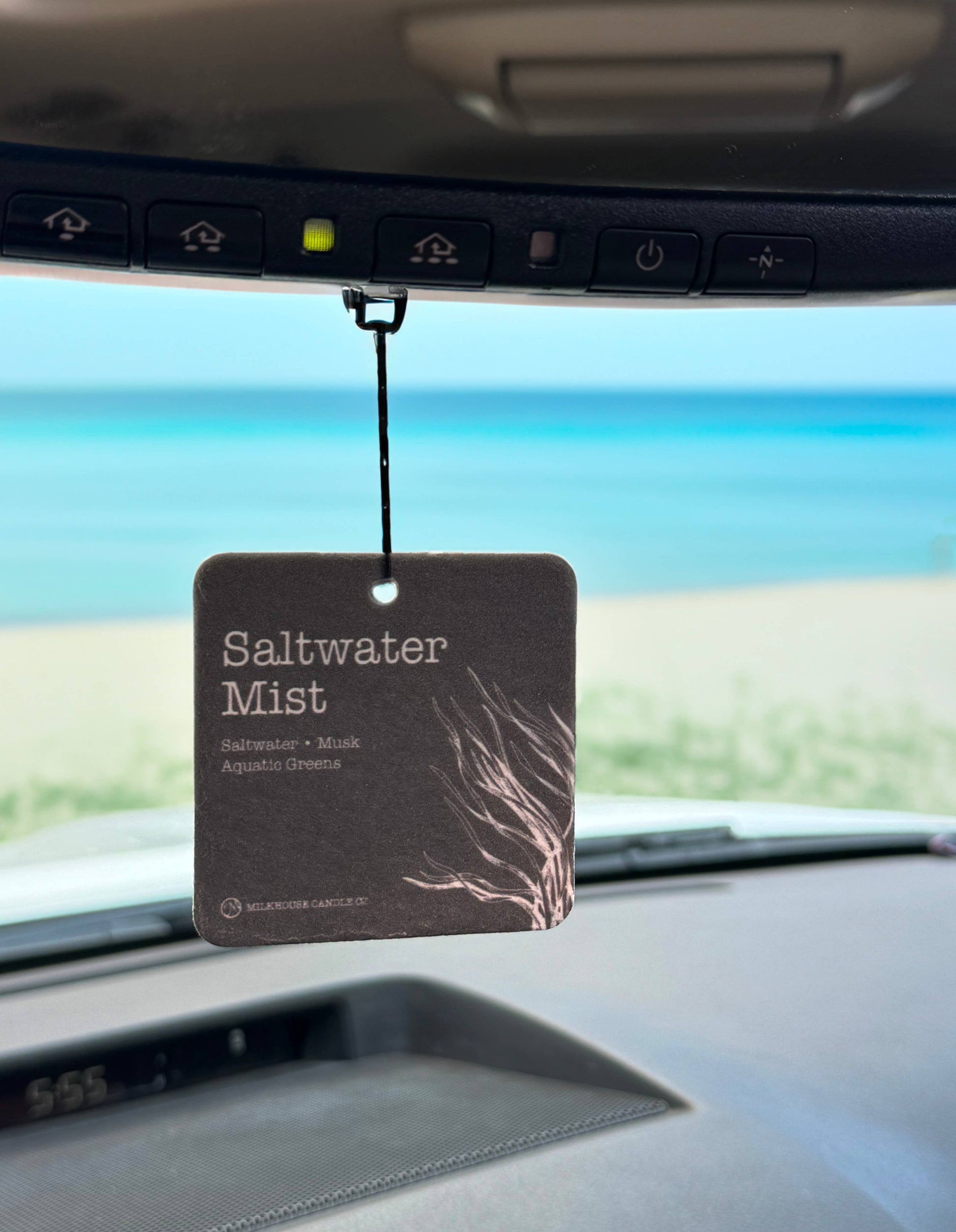 Car air freshener labeled 'Saltwater Mist' hanging from a car's sun visor with a beach view outside.